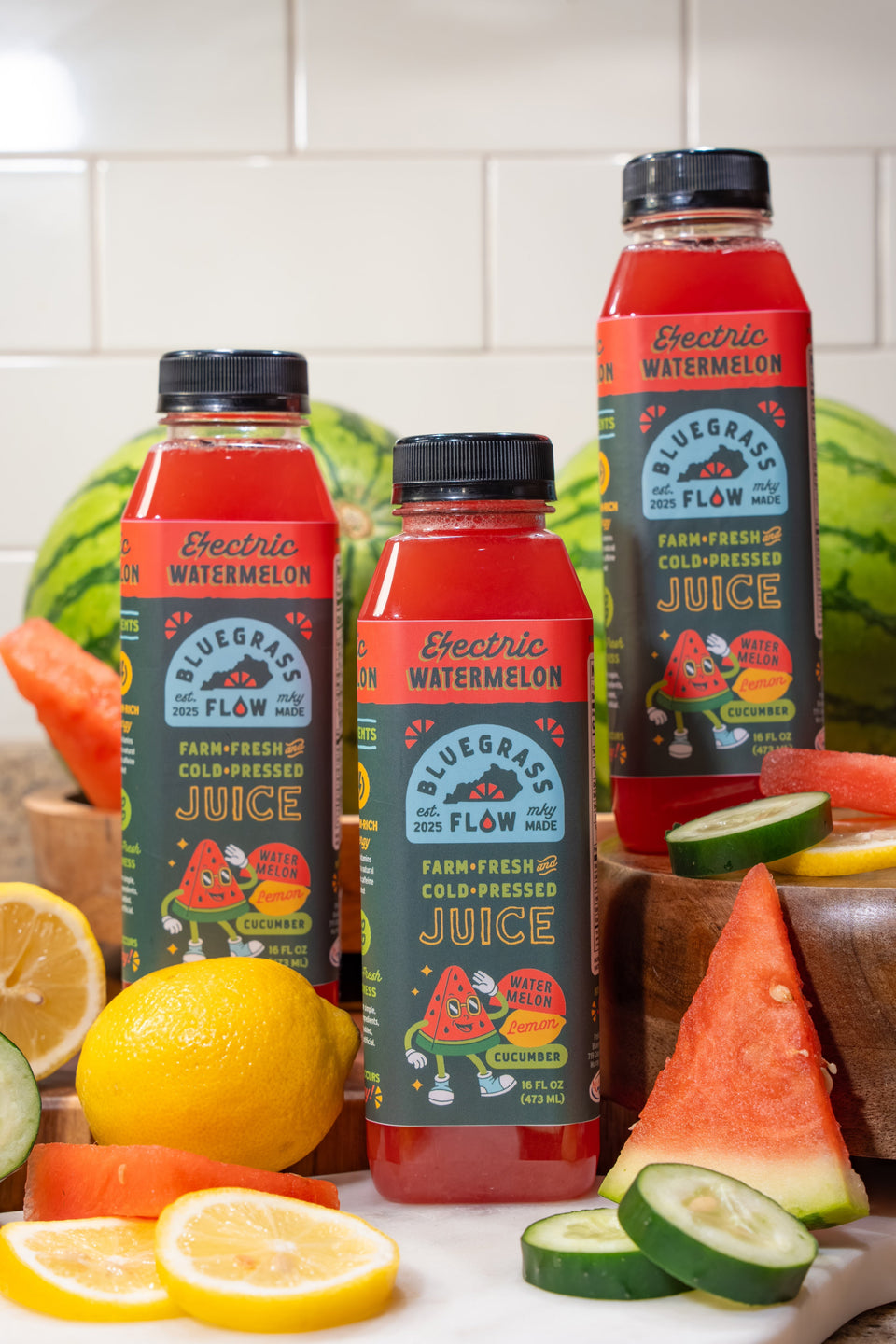 Bottles of Bluegrass Flow Juice with watermelon and lemon slices on a kitchen counter.